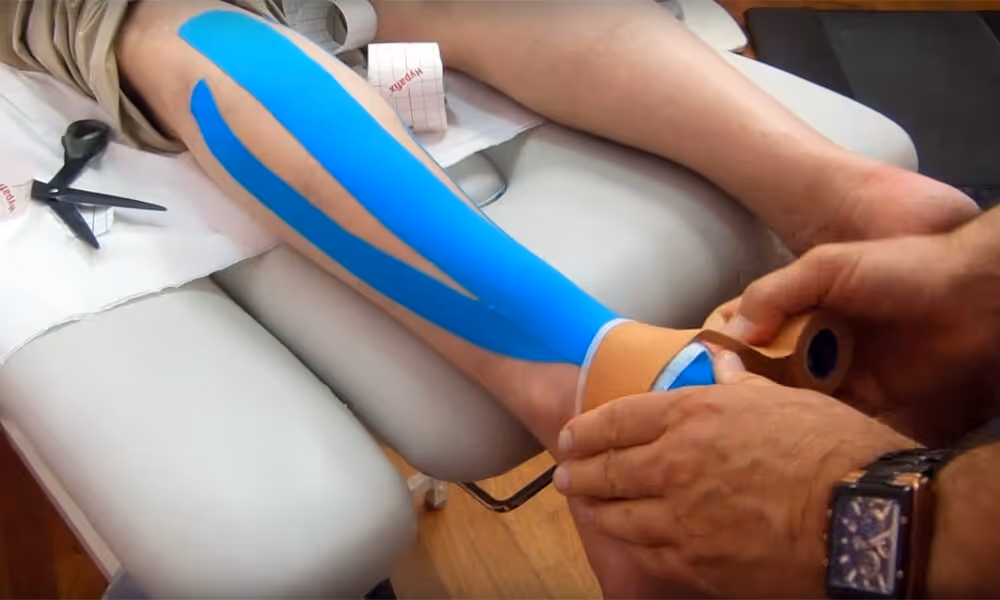 Person receiving blue kinesiology tape on their lower leg and ankle while sitting on a medical table.